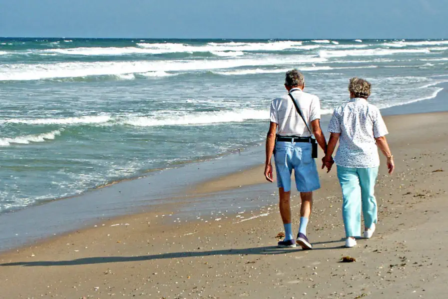 An elderly couple walk along a sandy beach on a hot day.